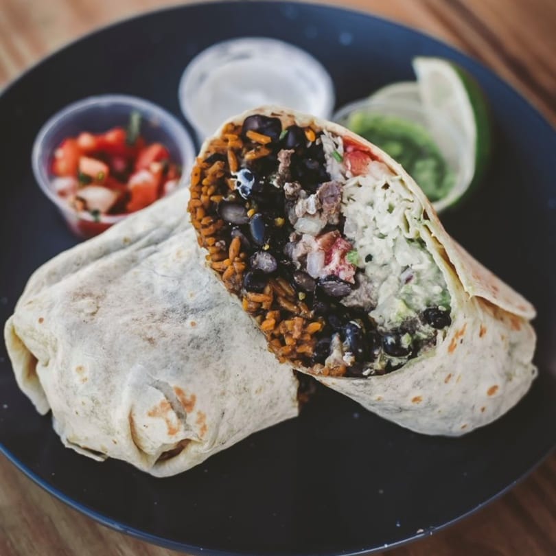 A large, cut burrito filled with rice, black beans, meat, and white sauce, with sides of salsa and guacamole.