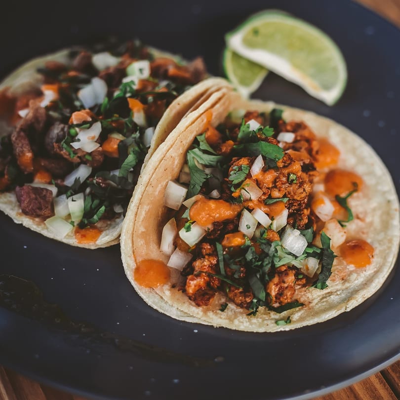 Two Azul Tacos, one with red meat and one with brown, topped with onions, cilantro, and sauce.