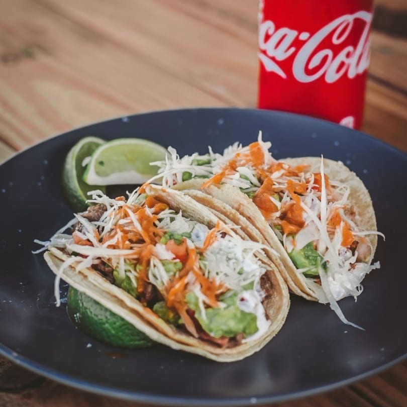 Two Azul Tacos with shredded cabbage, orange sauce, and guacamole, served with lime wedges and a Coca-Cola.